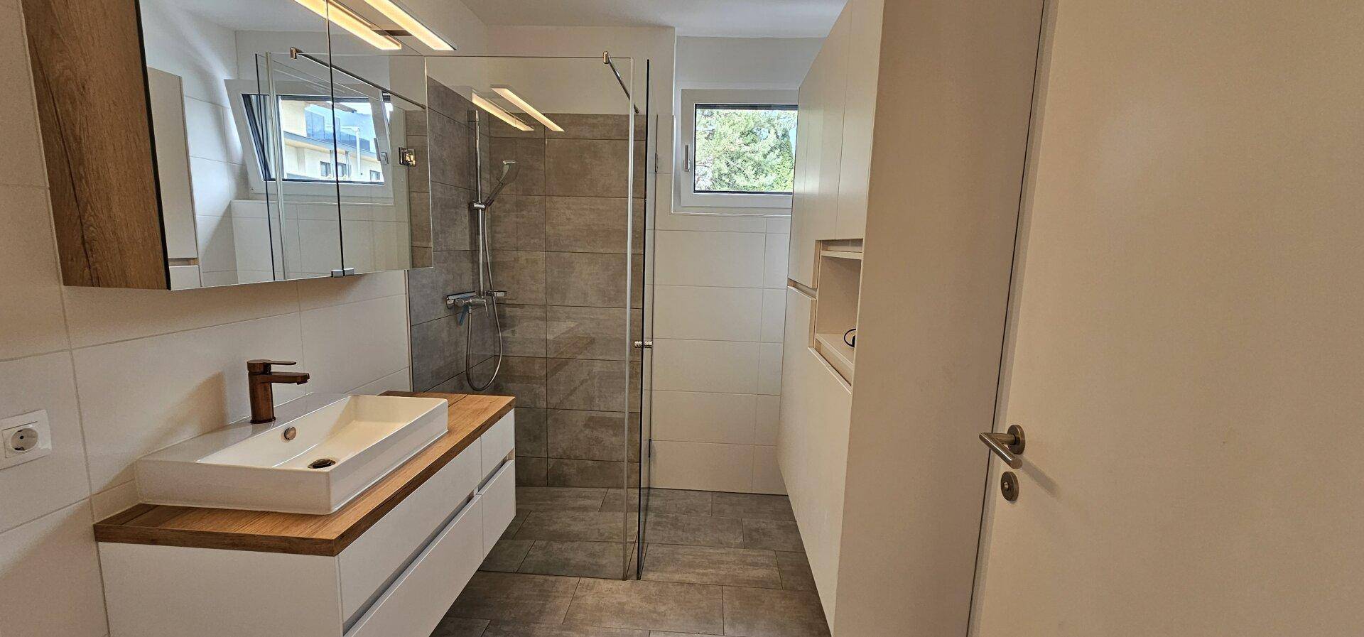 Modernes Badezimmer mit begehbarer Dusche, Holzwaschtisch und Fenster für Tageslichteinfall.