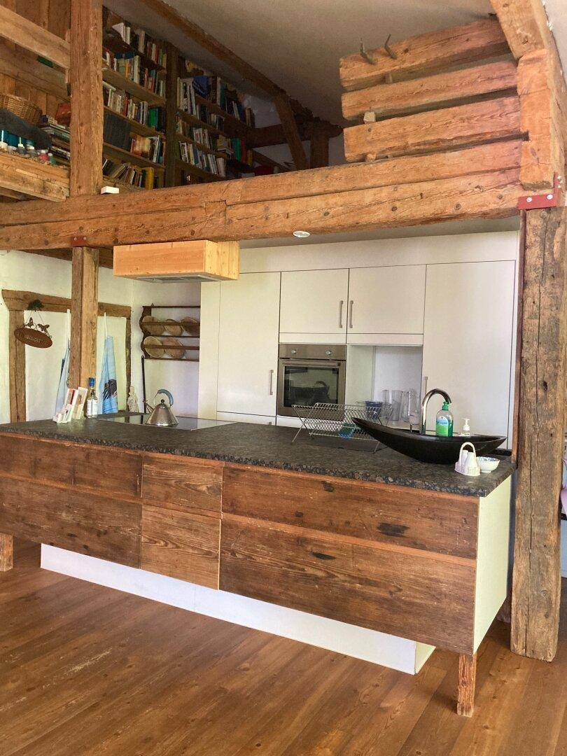 Offene Küche mit Kücheninsel und rustikalen Holzbalken, darüber ein Bücherregal im Loft-Stil.
