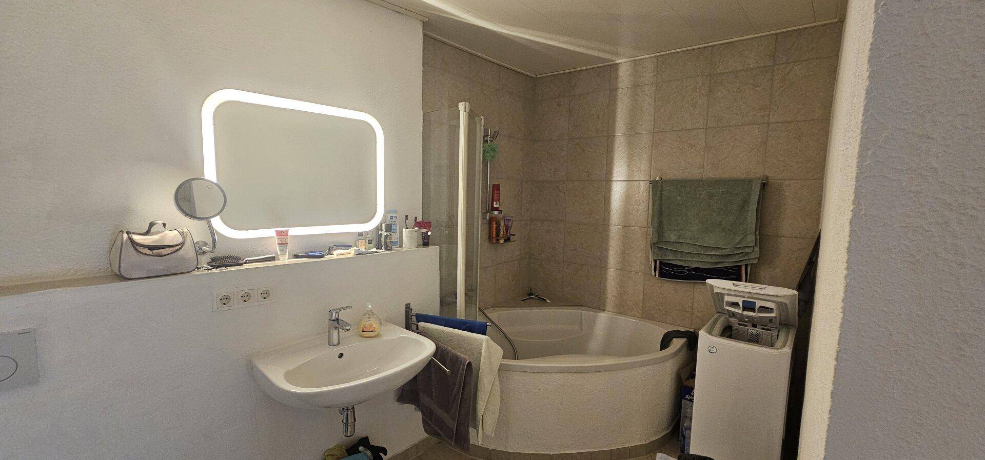 Funktionales Badezimmer mit Eckbadewanne, Waschbecken und Waschmaschine, hell beleuchtet.