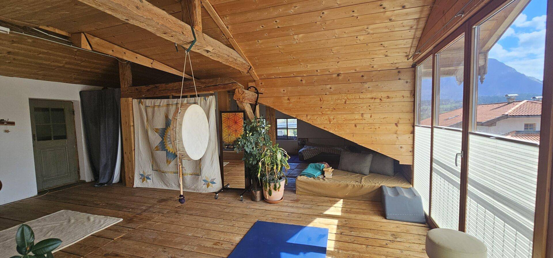 Geräumiger Dachgeschossraum mit Holzbalken, großen Fenstern und Blick auf die Berge.