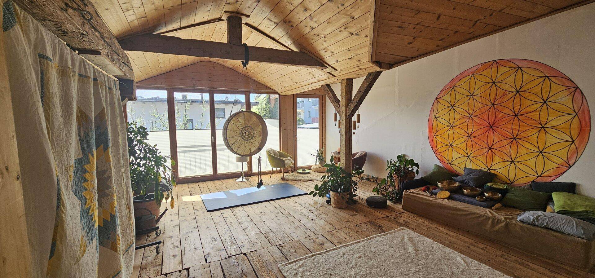 Heller Dachgeschossraum mit sichtbaren Holzbalken, großen Fenstern und spiritueller Dekoration.