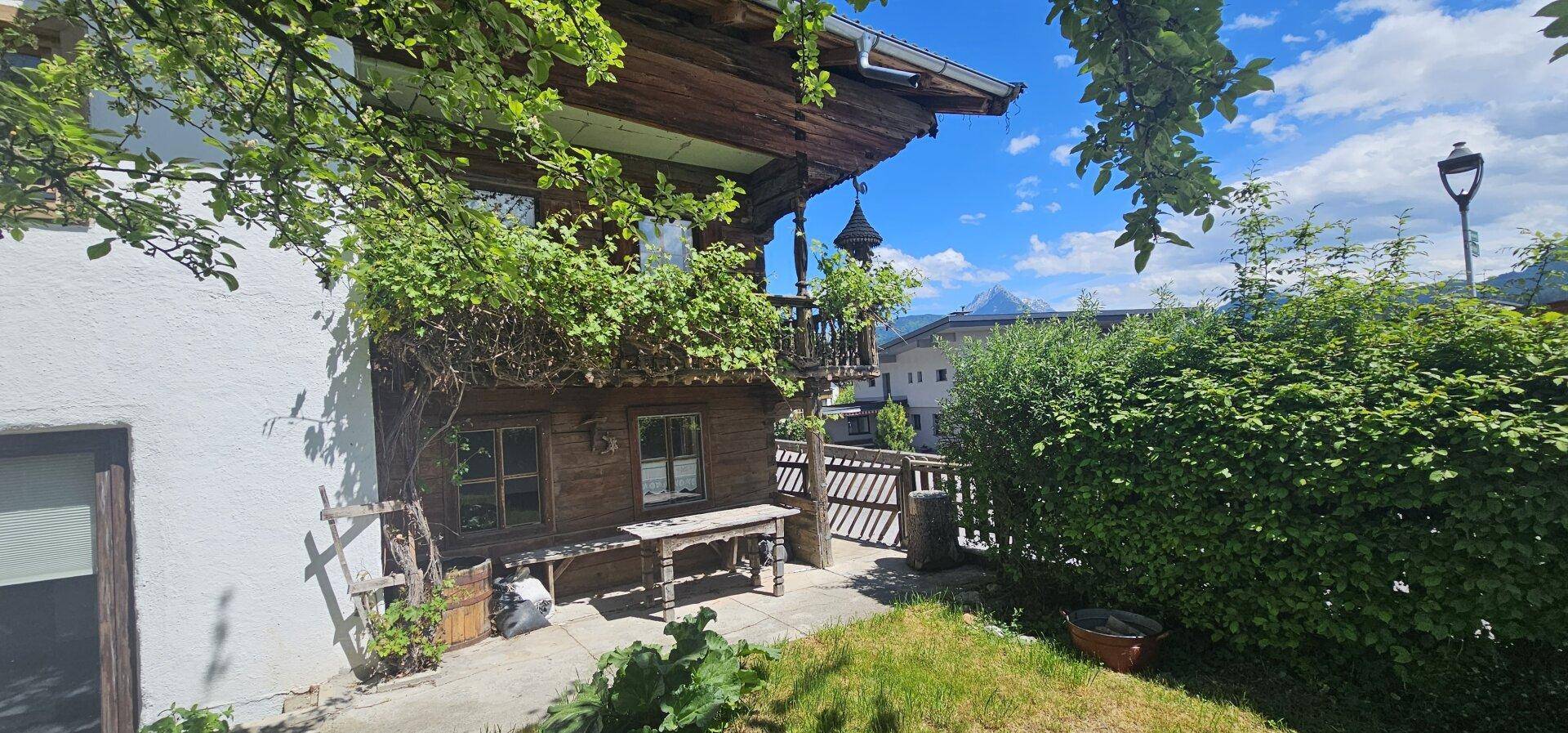 Traditionelles Holzhaus mit begrüntem Balkon und üppiger Vegetation im Außenbereich unter blauem Himmel.