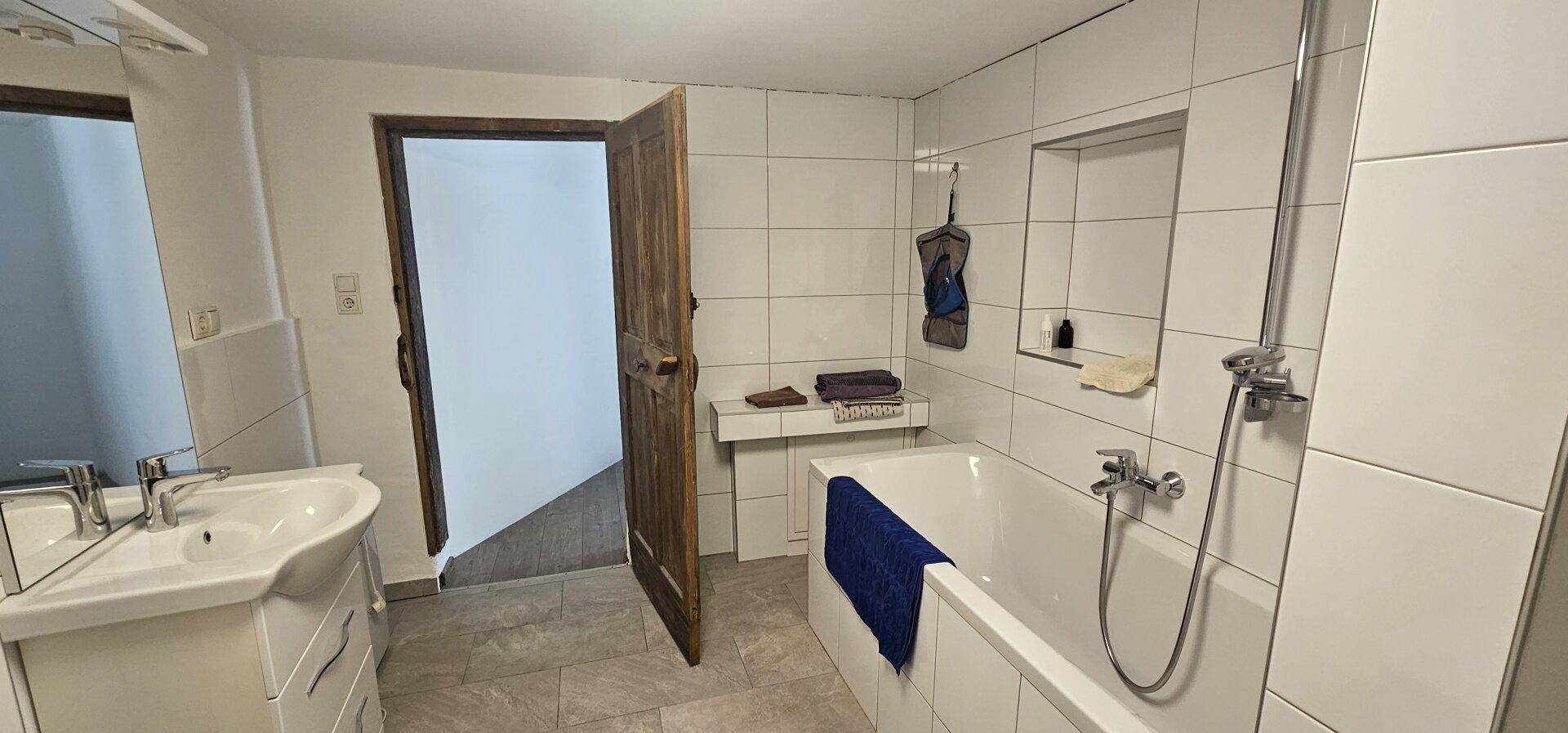 Helles Badezimmer mit weißen Fliesen, Badewanne, Waschbecken und einem Spiegel über dem Waschtisch.