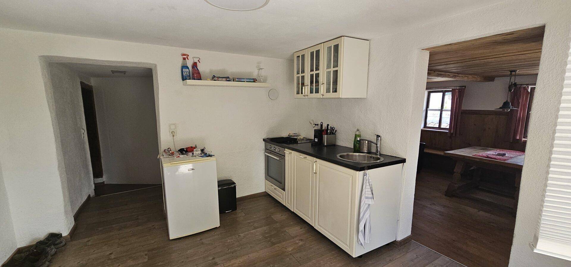 Kleine Küche mit weißen Schränken, Spüle, Herd und Kühlschrank, offen zum angrenzenden Essbereich.
