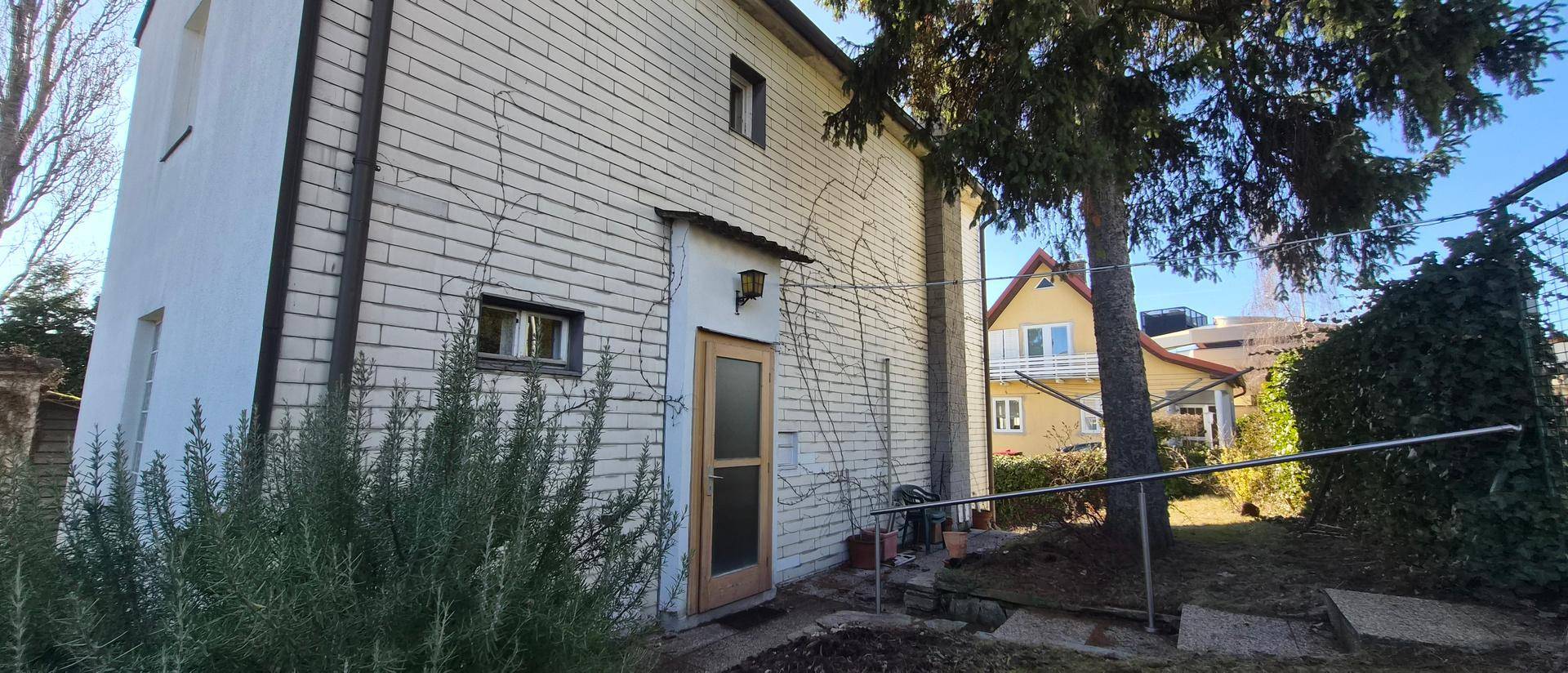Seitenansicht des Hauses mit Eingangstür und Garten, umgeben von Bäumen und Sträuchern.