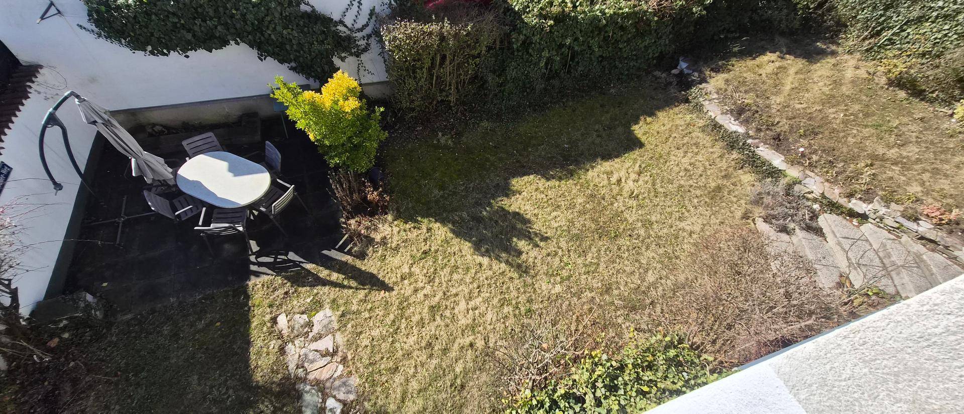 Vogelperspektive auf den Garten mit Rasenfläche und einem möblierten Sitzbereich unter einem Sonnenschirm.