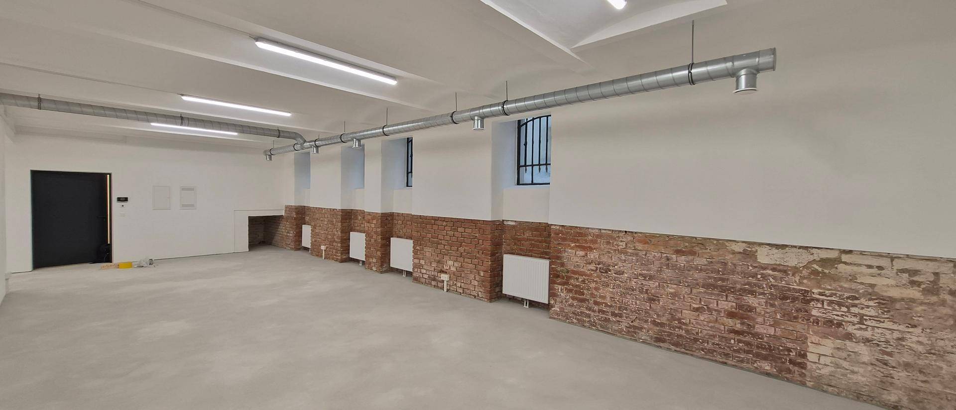 Heller, langer Raum mit weiß gestrichenen Wänden, unverputzten Ziegeln und einer schwarzen Tür am Ende.
