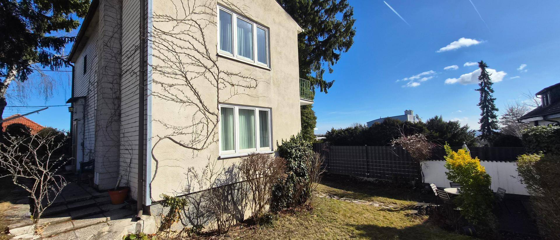 Seitenansicht des Hauses mit Garten und Hecken, umgeben von Bäumen an einem sonnigen Tag.