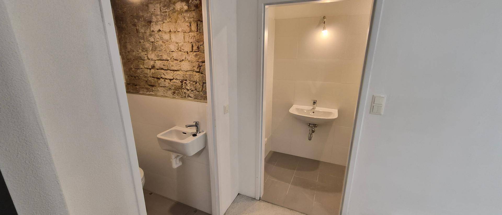 Zwei separate Toilettenbereiche mit Waschbecken, einer davon mit unverputzter Ziegelwand.