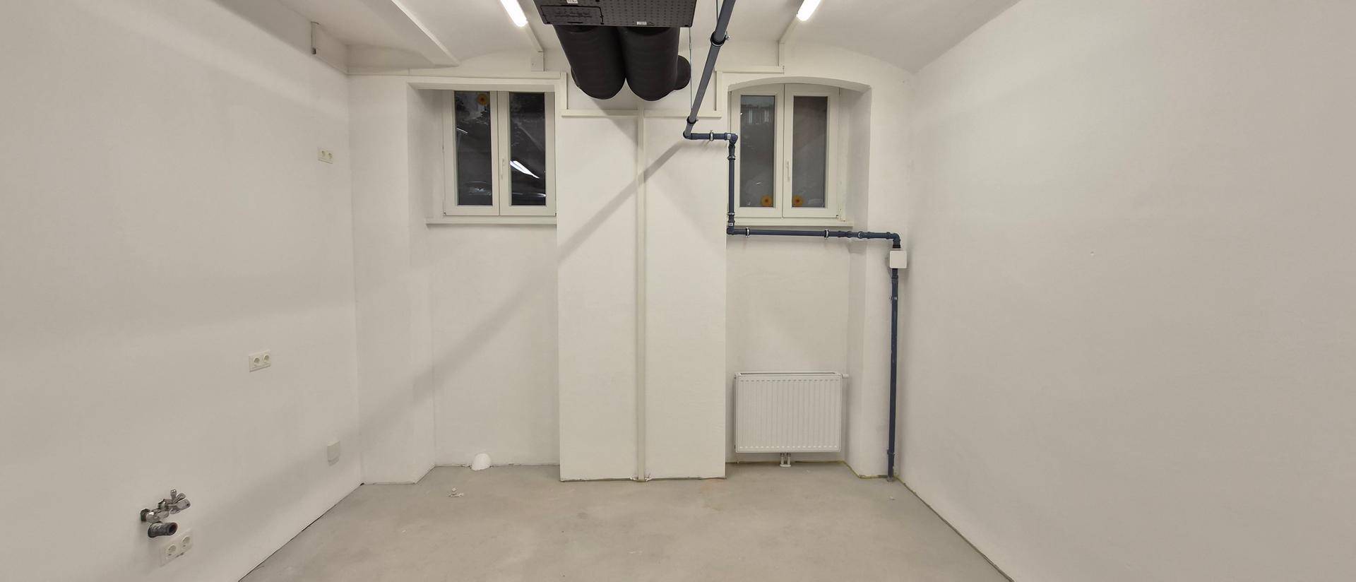 Kleinerer Raum mit zwei Fenstern, Heizkörper und sichtbaren Rohrleitungen an den Wänden.