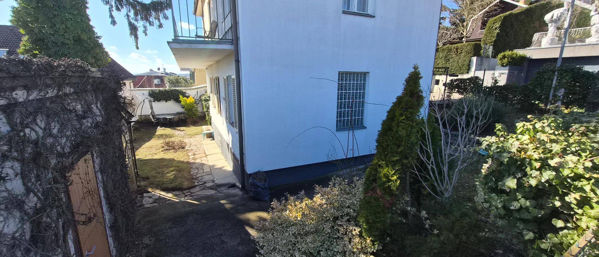 Schmaler Gartenweg entlang der Hauswand, umgeben von üppiger Vegetation und Bäumen.