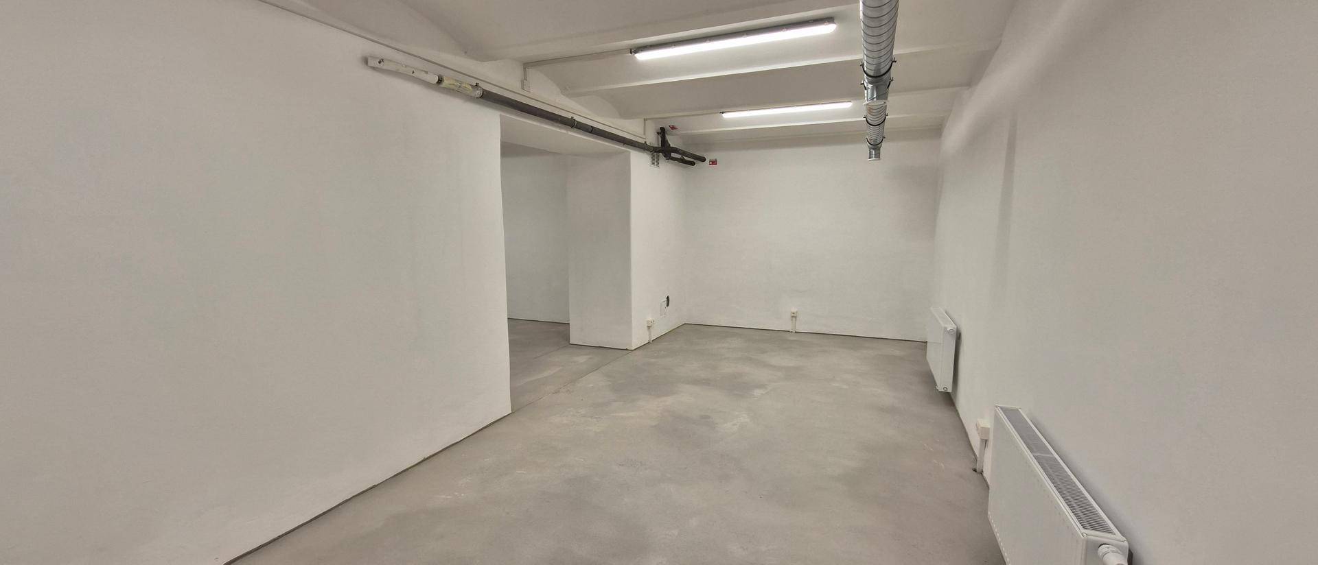 Schmalerer Bereich eines großen Raumes mit weißen Wänden, Betonboden und Heizkörpern.