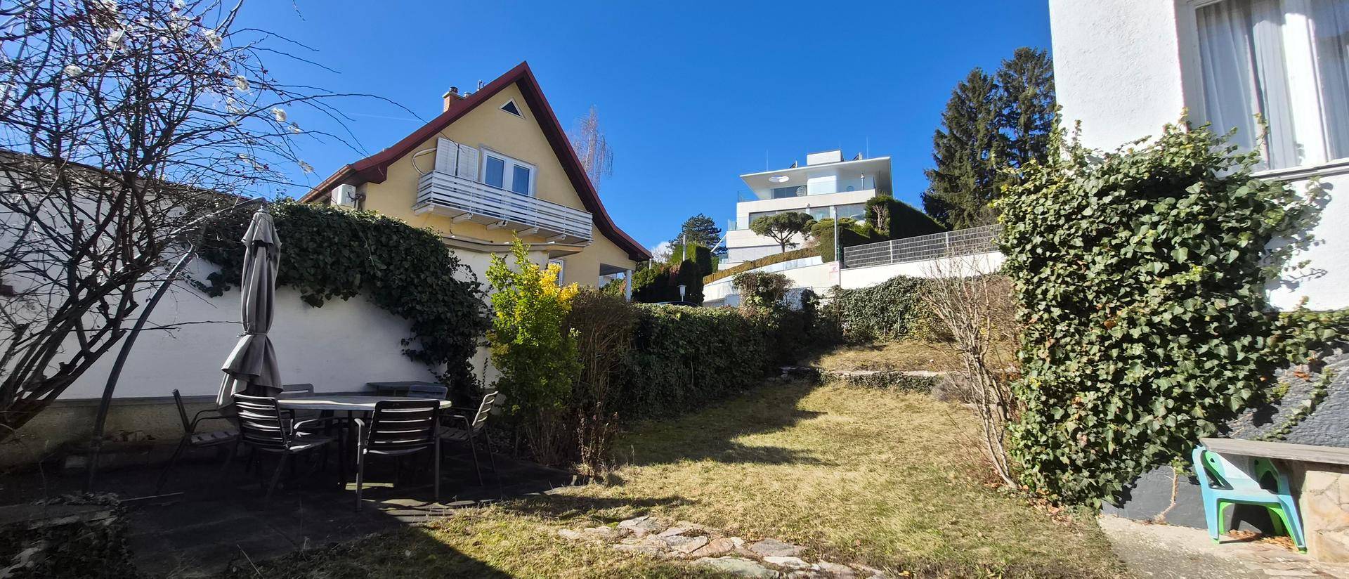 Gartenbereich mit Esstisch und Sonnenschirm, umgeben von Hecken und Blick auf Nachbarhäuser.