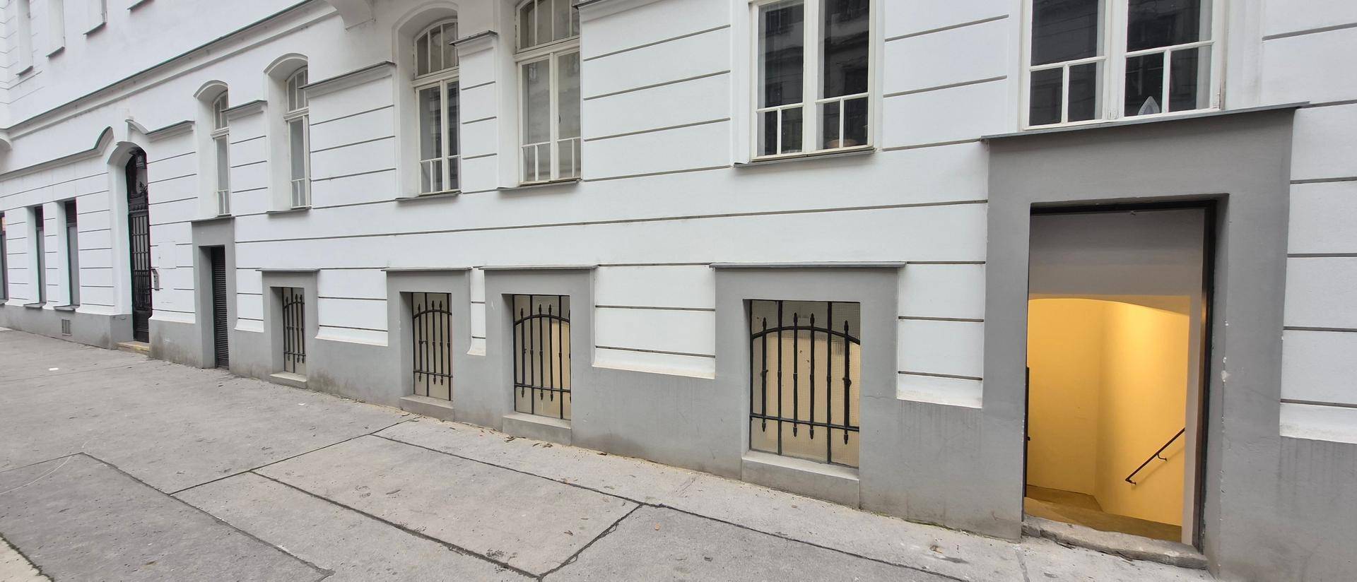 Außenansicht eines Gebäudes mit Eingang zum Untergeschoss und vergitterten Fenstern auf Straßenniveau.