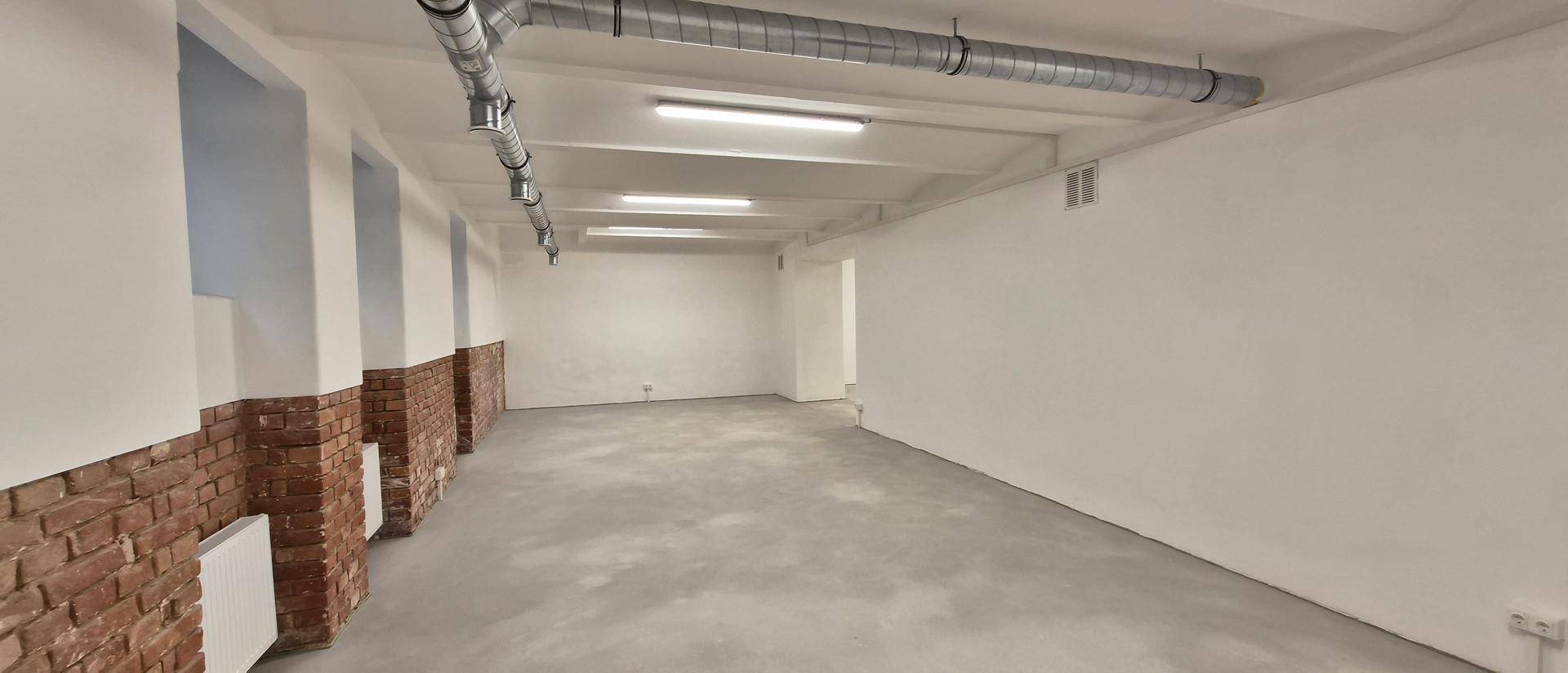 Weiter Blick in einen leeren Raum mit hohen Decken, Betonboden und sichtbaren Lüftungsrohren.