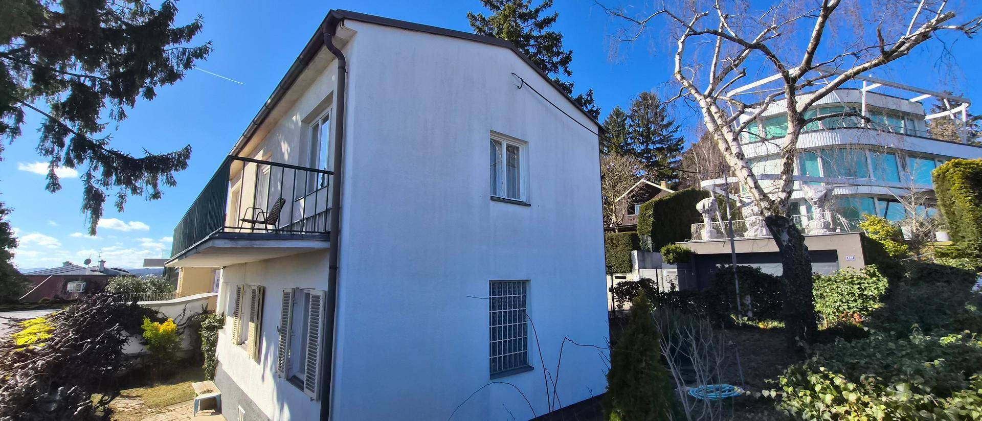 Rückansicht des Hauses mit Balkon und Blick auf benachbarte moderne Gebäude und grüne Umgebung.