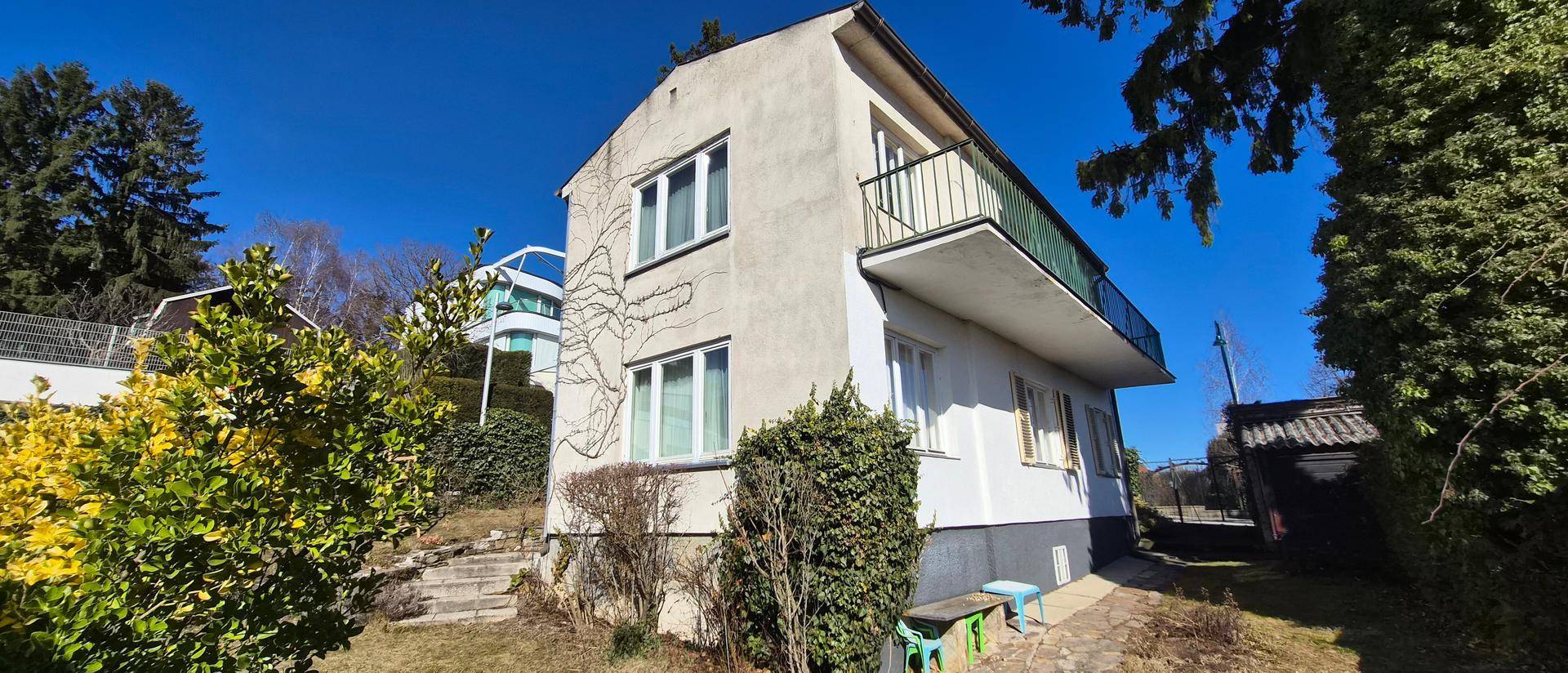 Ansicht des Hauses mit einem Balkon und umliegender Vegetation unter strahlend blauem Himmel.