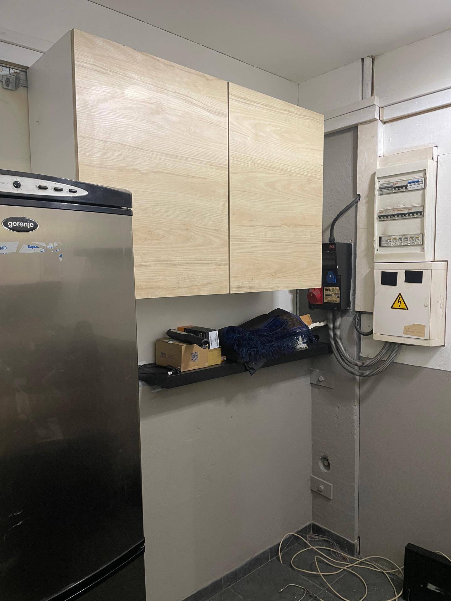 Kleiner Hauswirtschaftsraum mit einem Kühlschrank, Holzschränken und einem sichtbaren elektrischen Sicherungskasten.