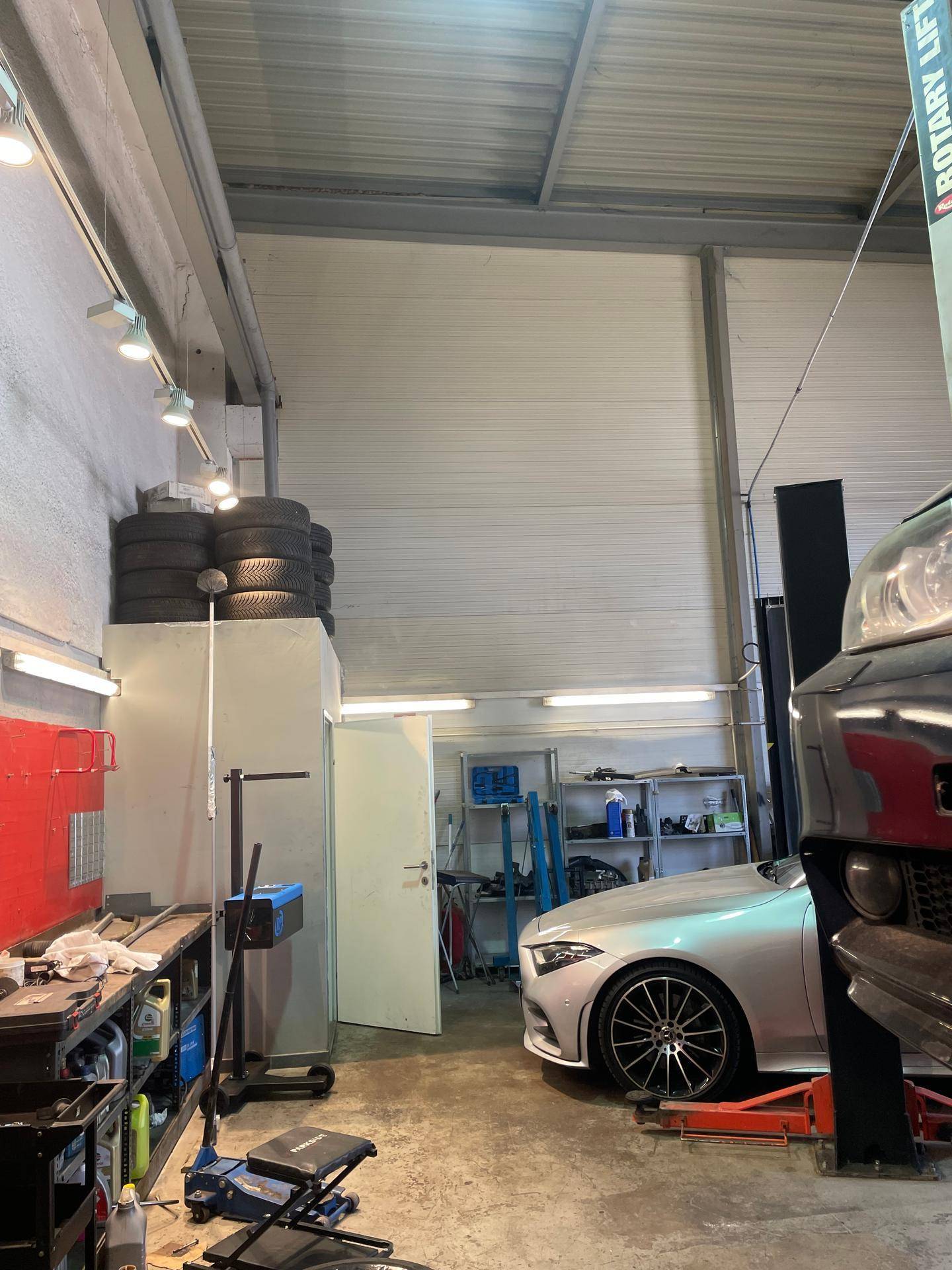 Ecke einer Autowerkstatt mit Werkzeugen, gestapelten Reifen und einem teilweise sichtbaren Fahrzeug auf dem Betonboden.