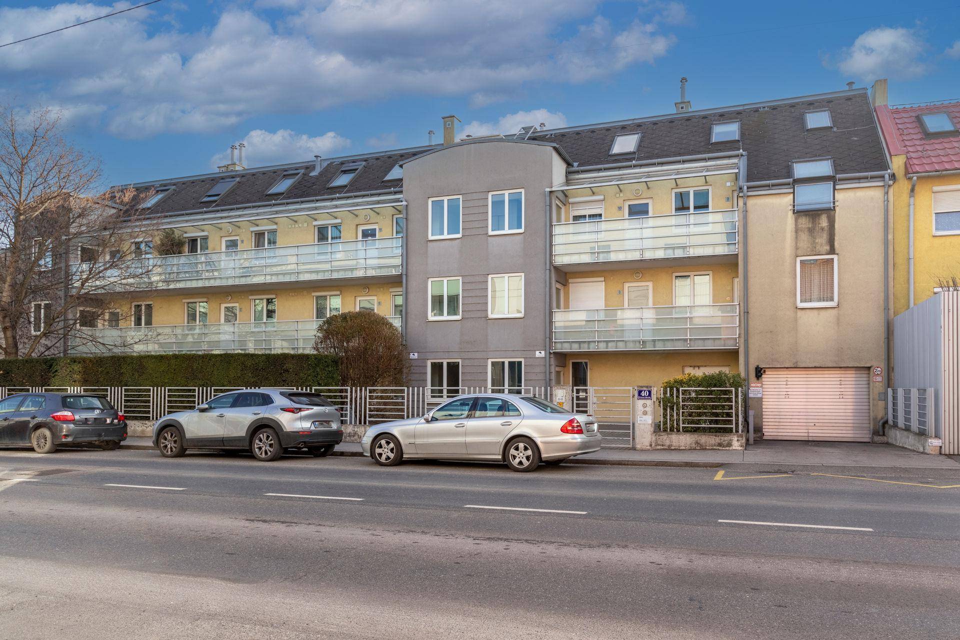 Mehrfamilienhaus mit Balkonen und Parkplätzen an der Straße, unter blauem Himmel.