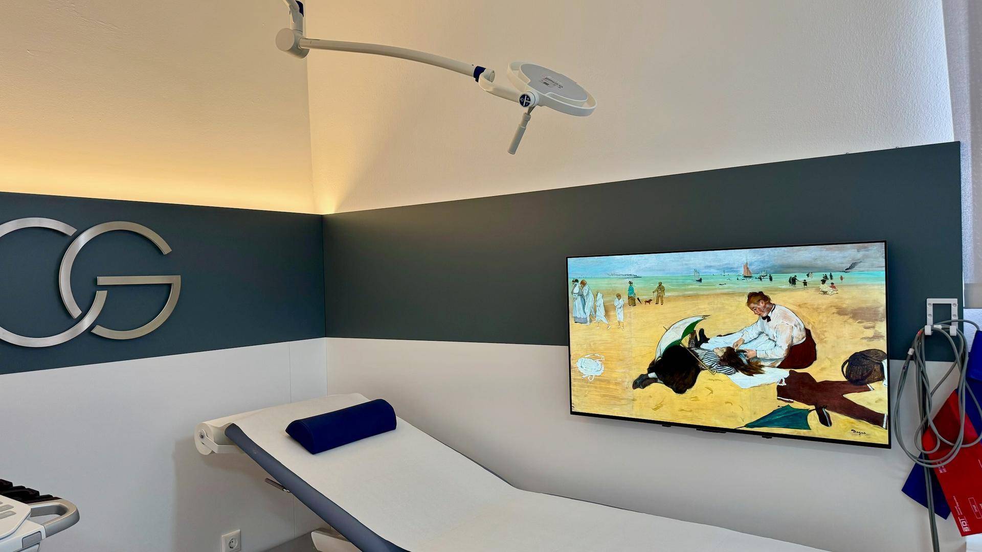 Untersuchungsraum mit einer Liege, einem Wandfernseher und medizinischer Beleuchtung an der Decke.