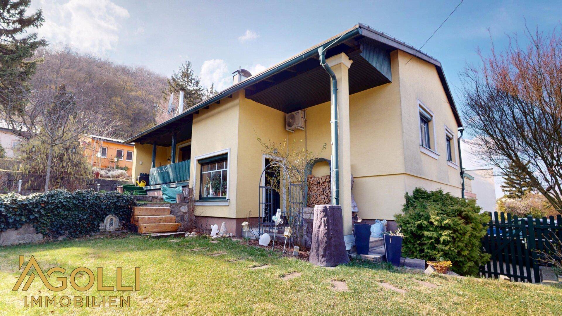 Seitenansicht des Hauses vom Garten aus mit überdachtem Eingangsbereich, Holzstapel und gepflegter Rasenfläche.