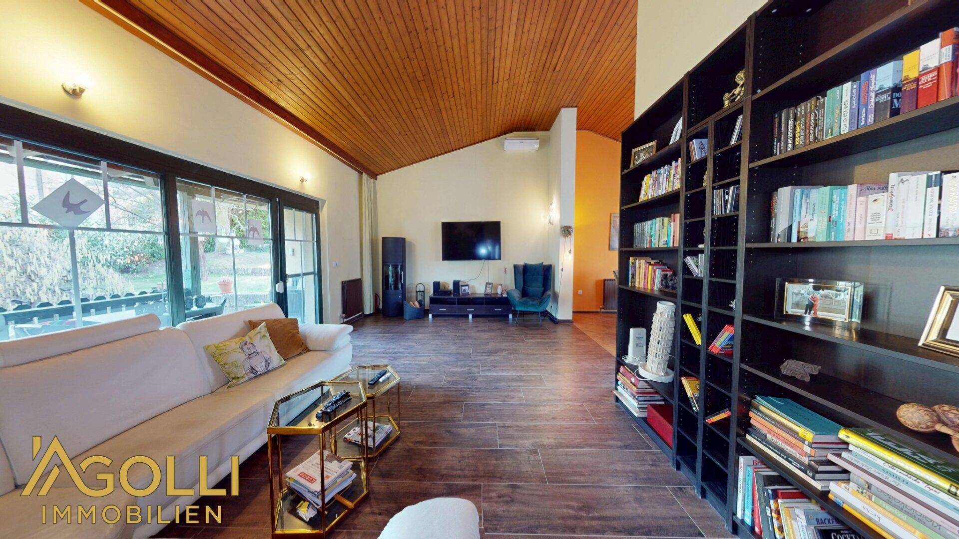 Geräumiges Wohnzimmer mit Holzbalkendecke, Dielenboden, großem Bücherregal und Kaminofen.