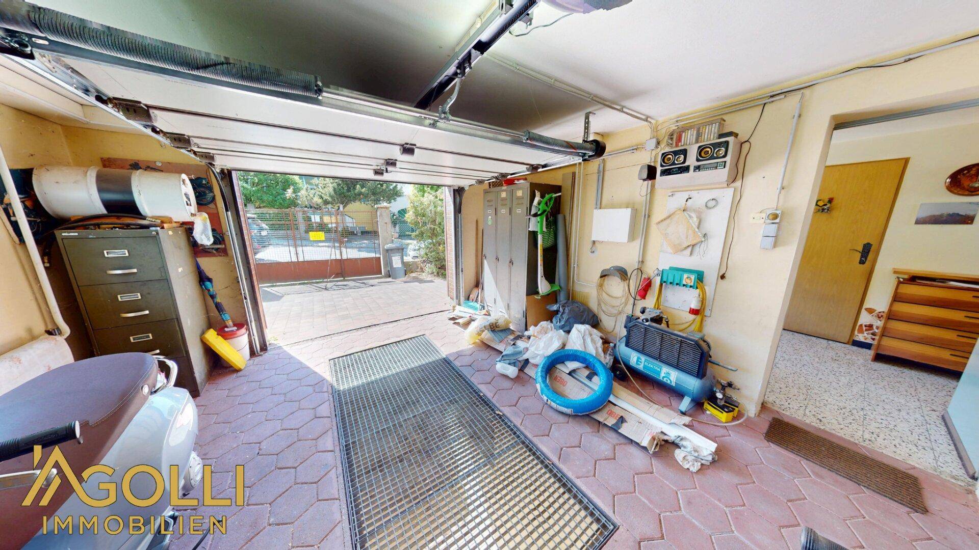 Geräumige Garage mit Werkzeugen, Lagerregalen und offenem Tor zur Einfahrt.