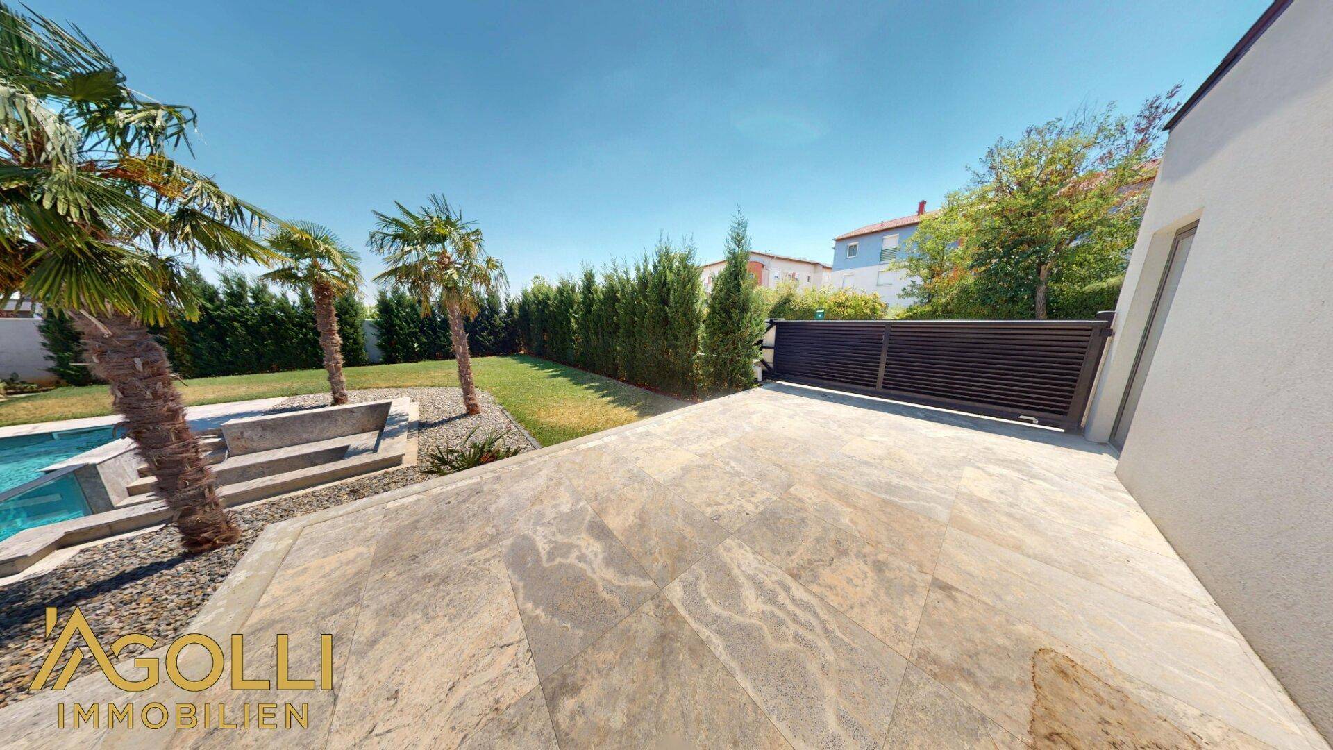 Gepflasterte Terrasse neben dem Poolbereich mit Palmen und einer hohen Hecke.