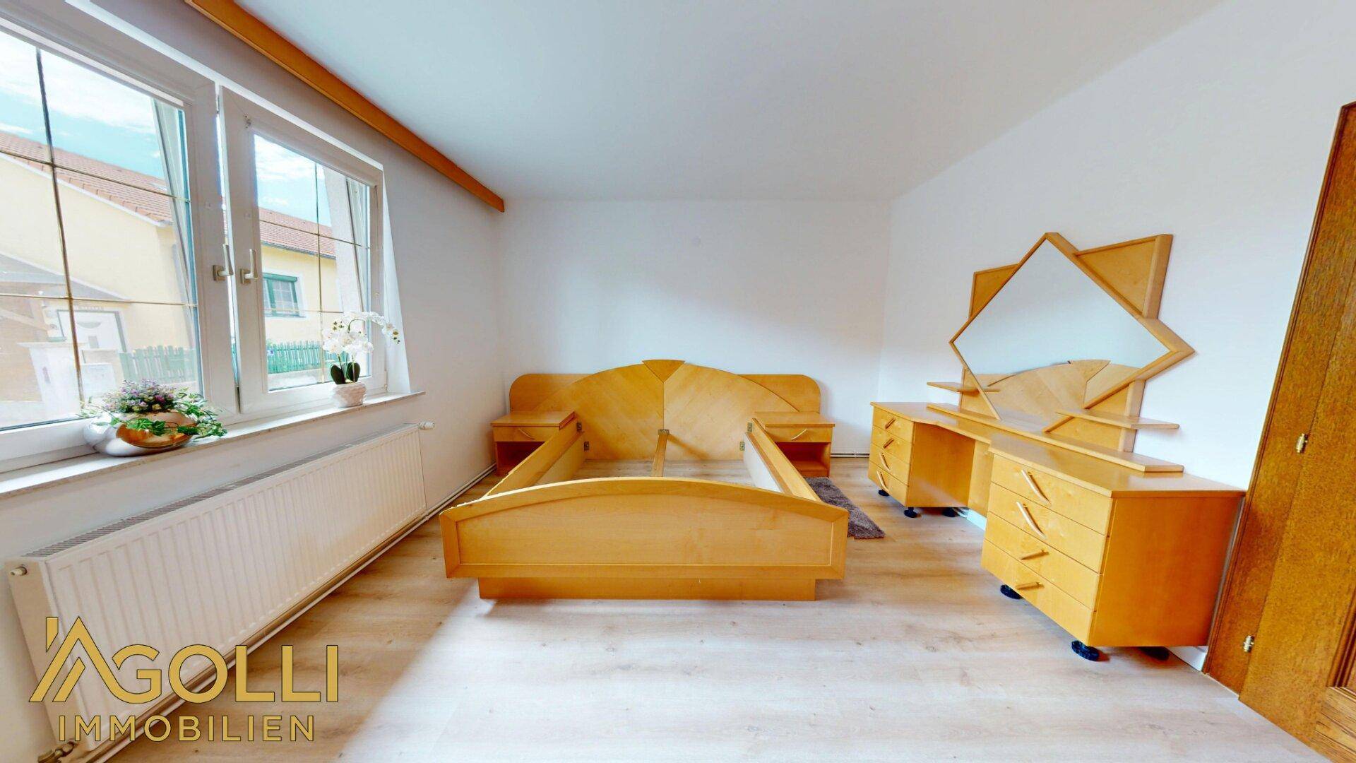 Schlafzimmer mit hellem Laminatboden, Holzmöbeln und großen Fenstern mit Blick nach draußen.