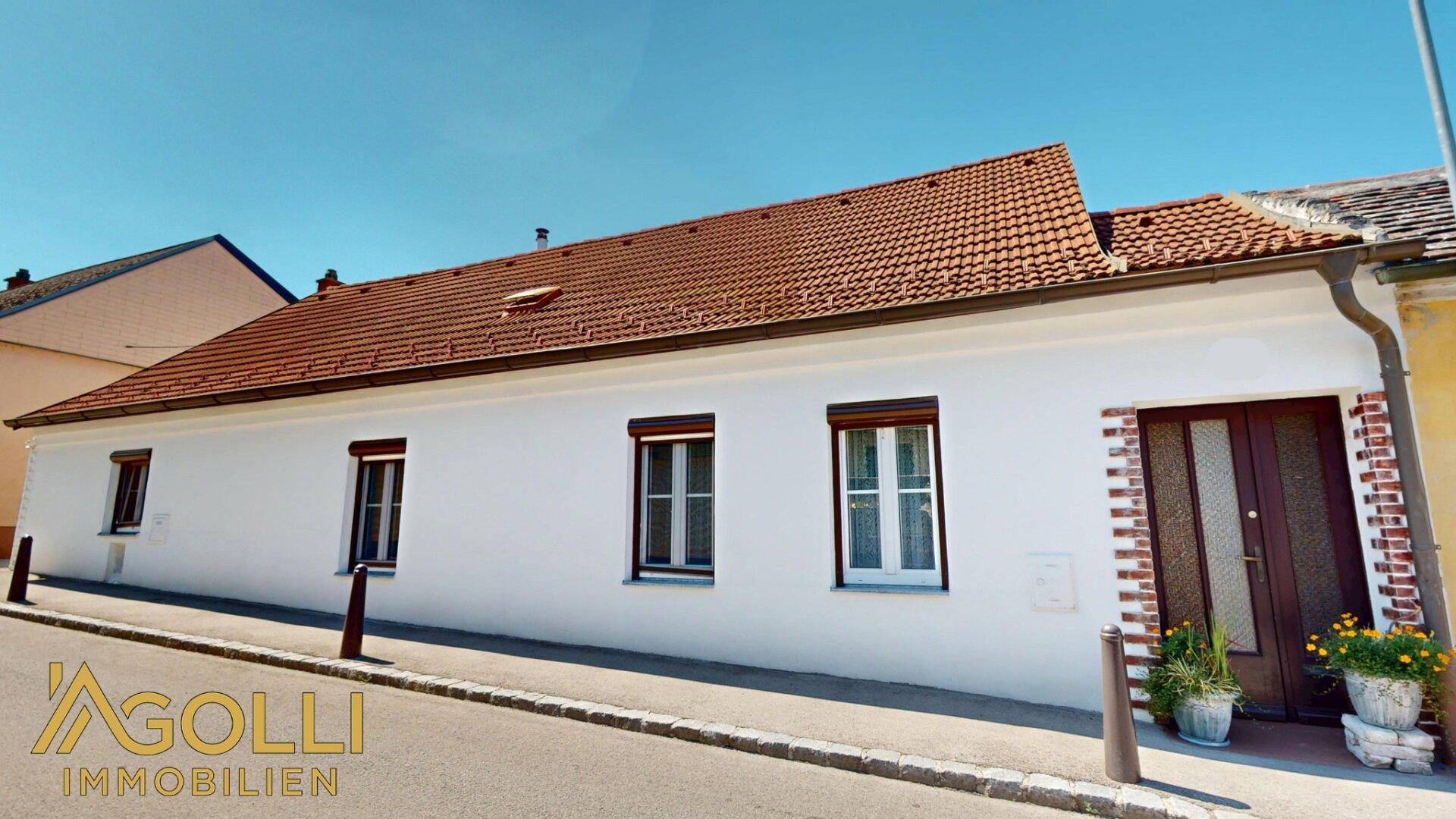 Weißes Haus mit Ziegeldach und braunen Fensterläden, direkt an der Straße gelegen.