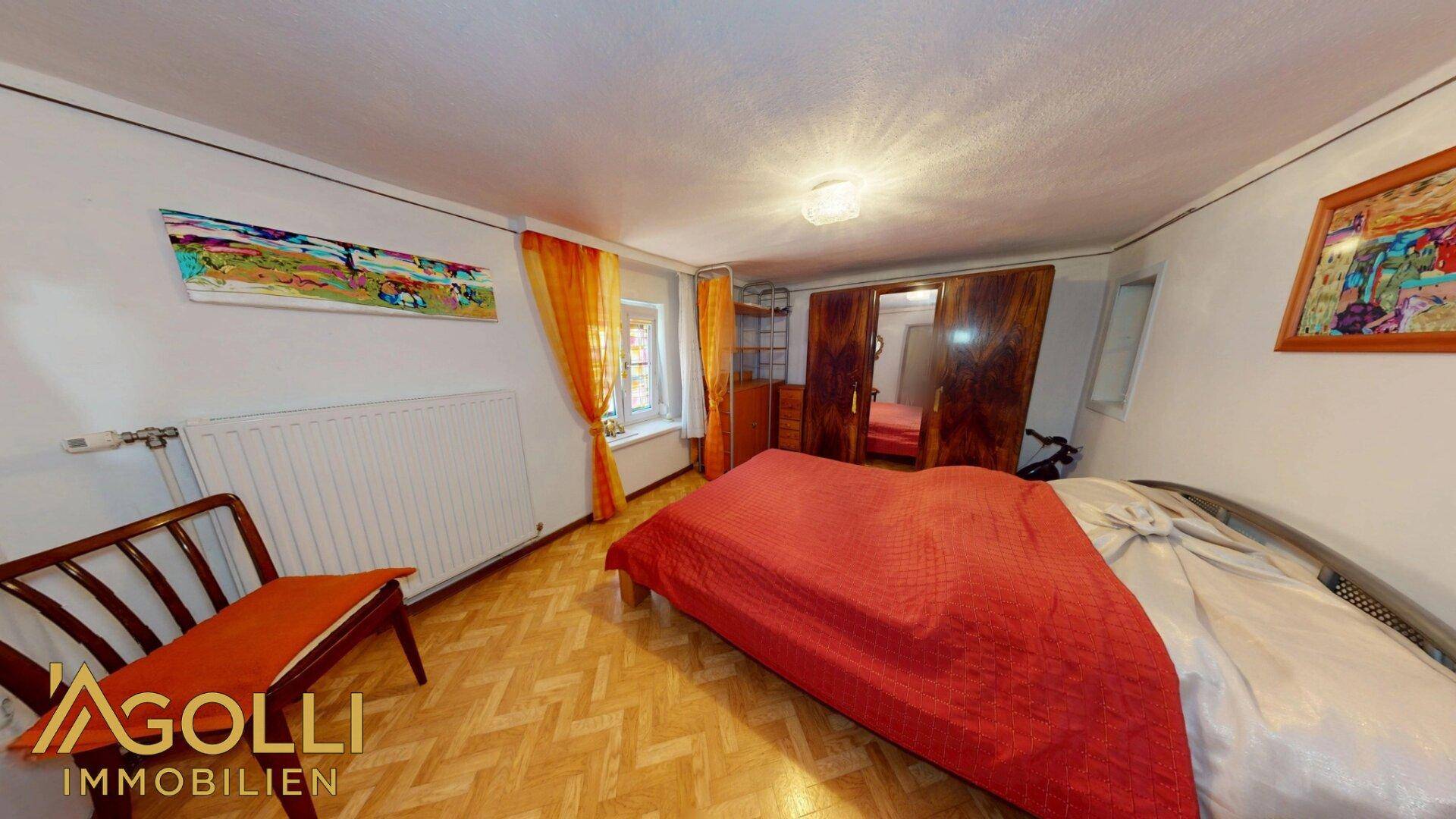 Gemütliches Schlafzimmer mit roter Bettdecke, Fischgrätparkett und großem Kleiderschrank.