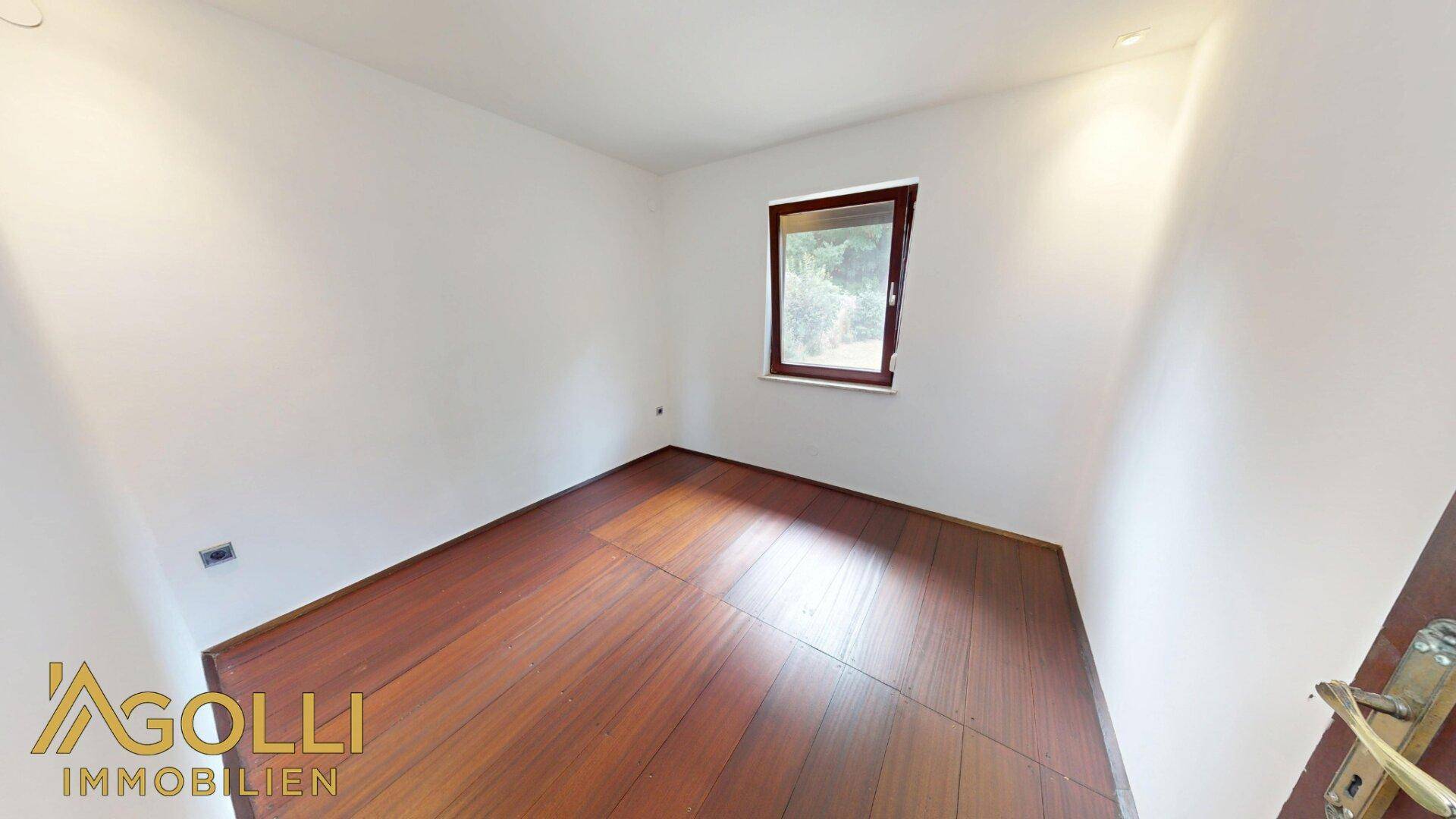 Leeres Schlafzimmer mit dunklem Holzboden und einem Fenster, das Tageslicht hereinlässt.