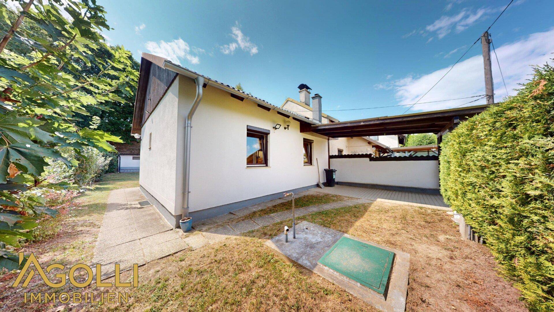 Seitenansicht des Hauses mit Carport, gepflastertem Bereich und Gartenfläche unter blauem Himmel.