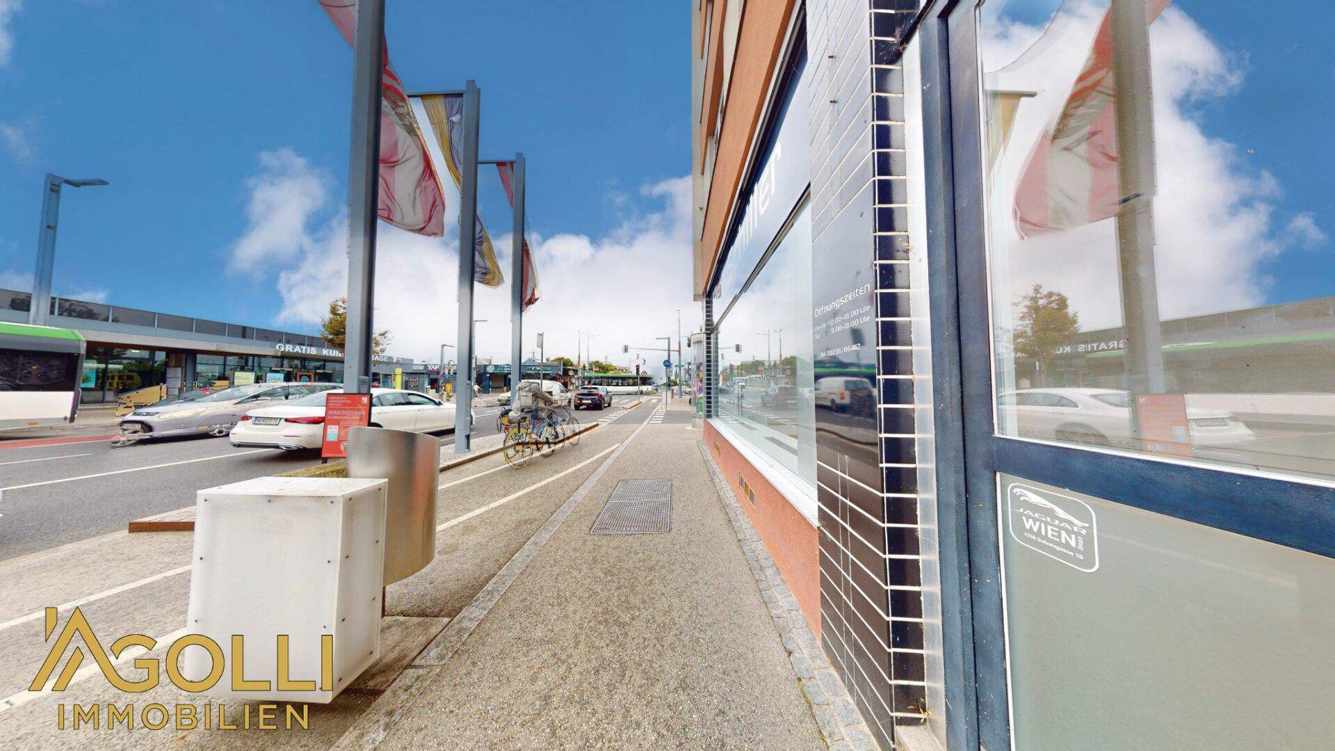 Belebte Straße vor dem Gebäude mit Bushaltestelle und vorbeifahrenden Fahrzeugen.