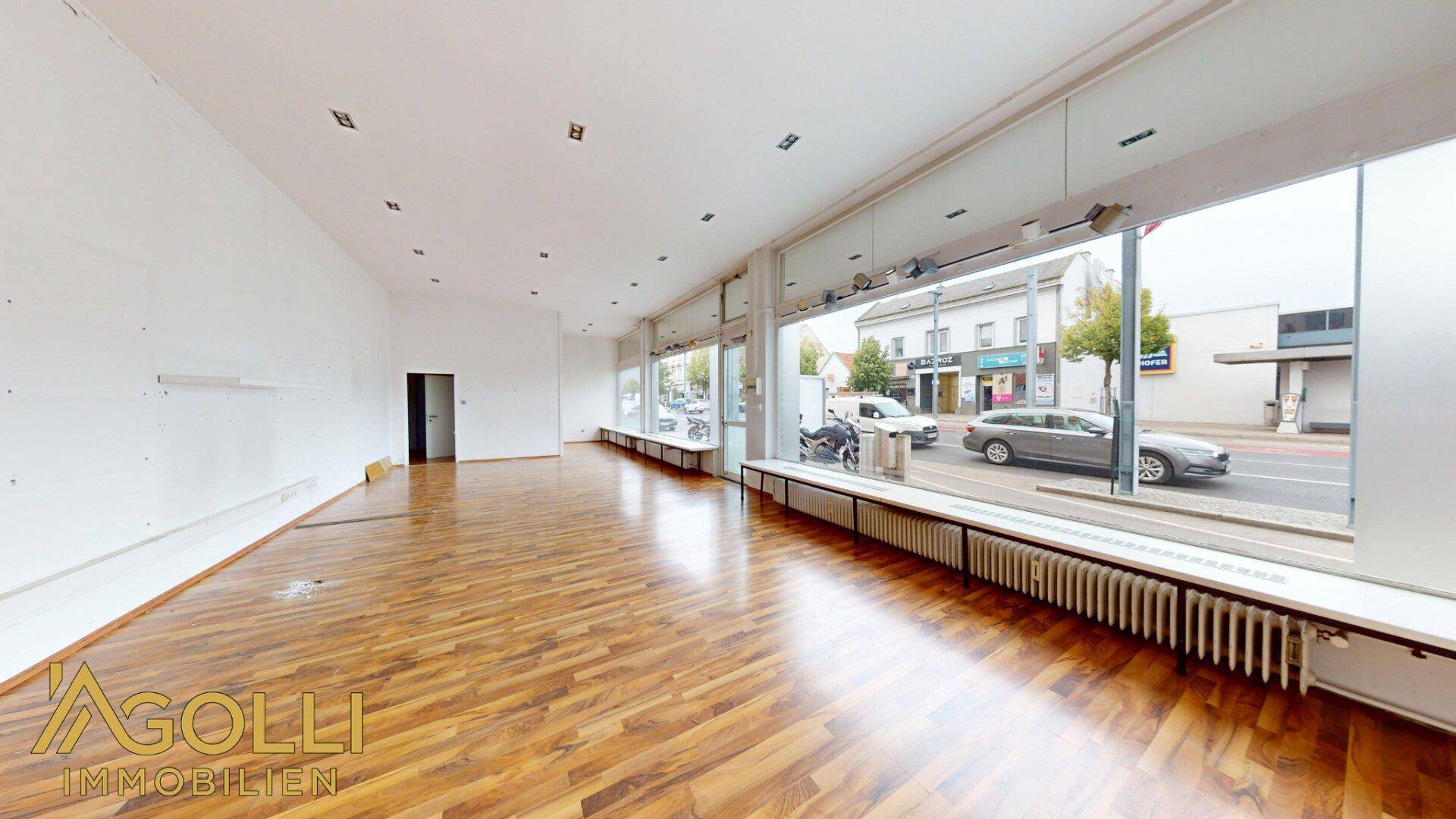 Langer Verkaufsraum mit durchgehender Fensterfront zur Straße und Heizkörpern.