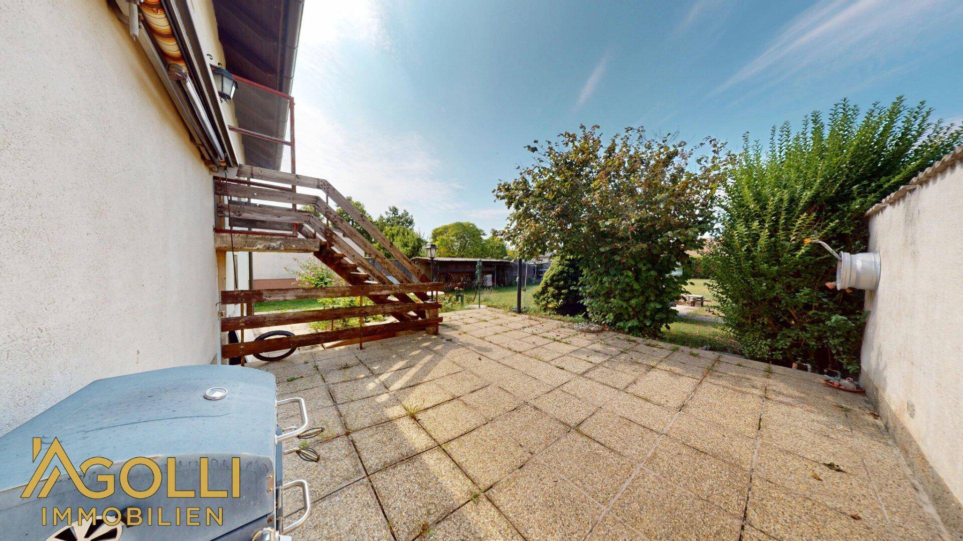 Gepflasterte Terrasse mit Grill und Zugang zum Garten über eine Holztreppe.