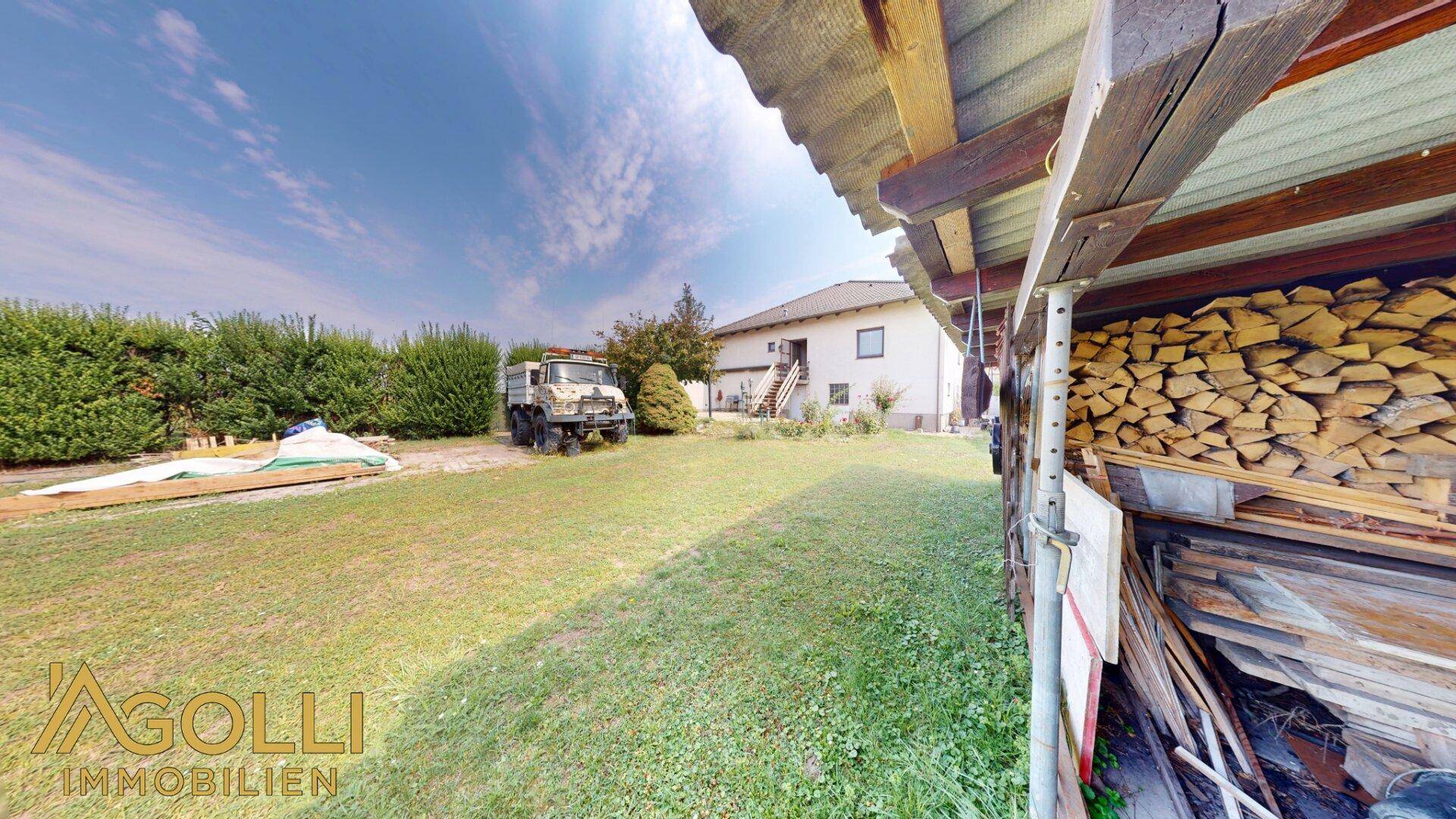 Weitläufiger Garten mit Holzstapel unter einem Unterstand und Blick auf die Rückseite des Hauses.