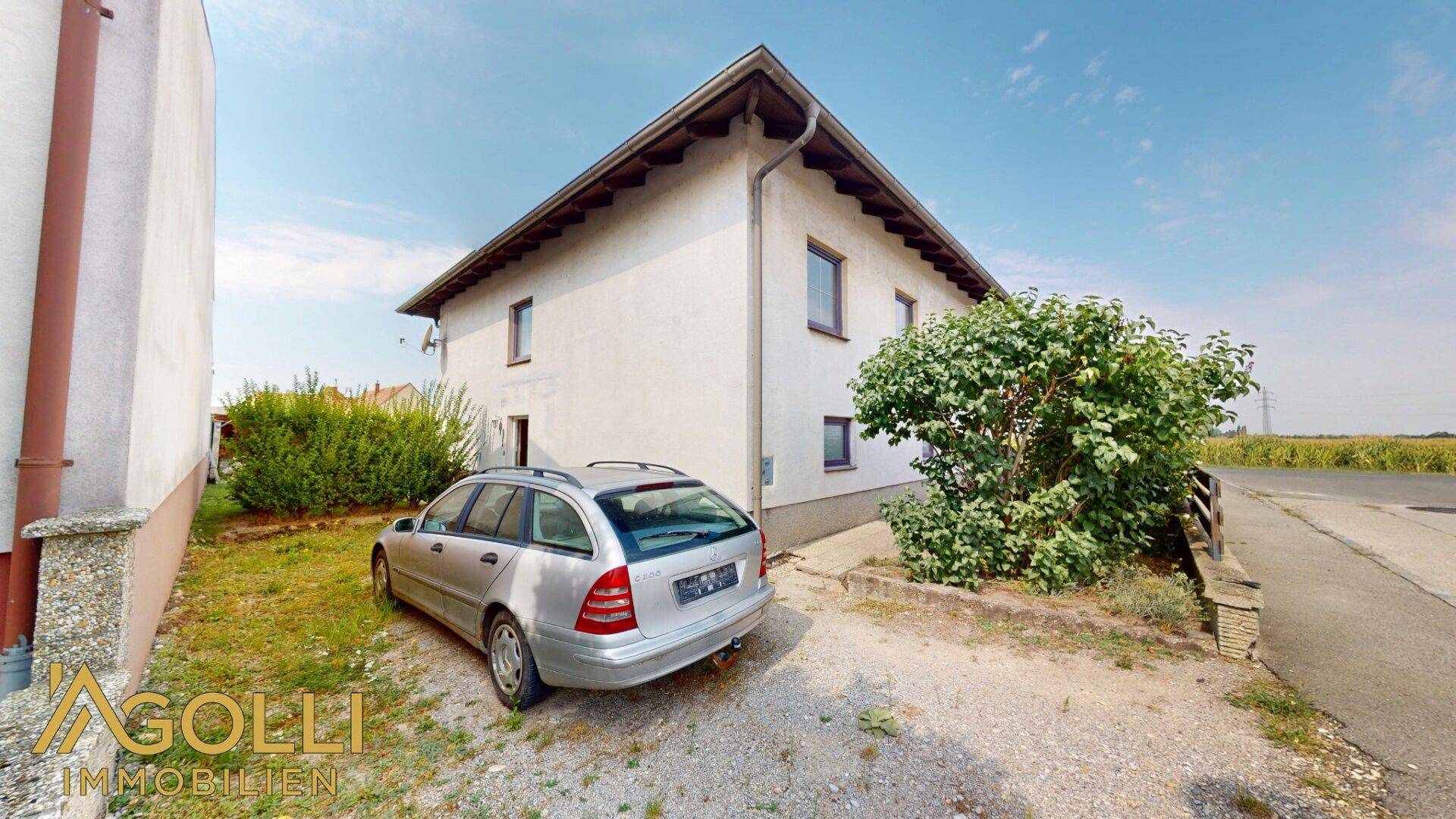 Seitenansicht des Hauses mit einem geparkten Auto auf dem Schotterweg und angrenzendem Feld.