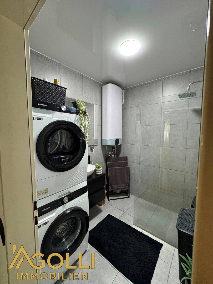 Modernes Badezimmer mit Dusche, Waschmaschine, Trockner und komplett grauen Fliesen.