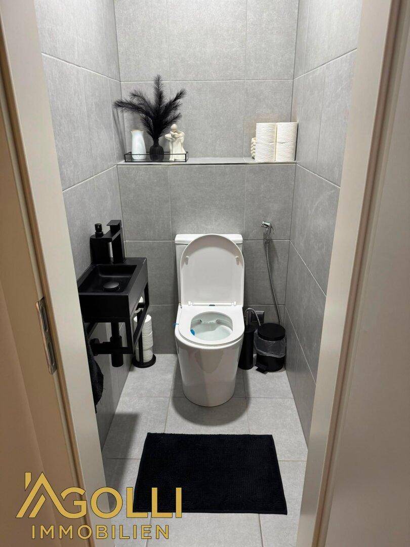 Kleines Gäste-WC mit modernen grauen Fliesen, wandmontierter Toilette und kleinem Waschbecken.
