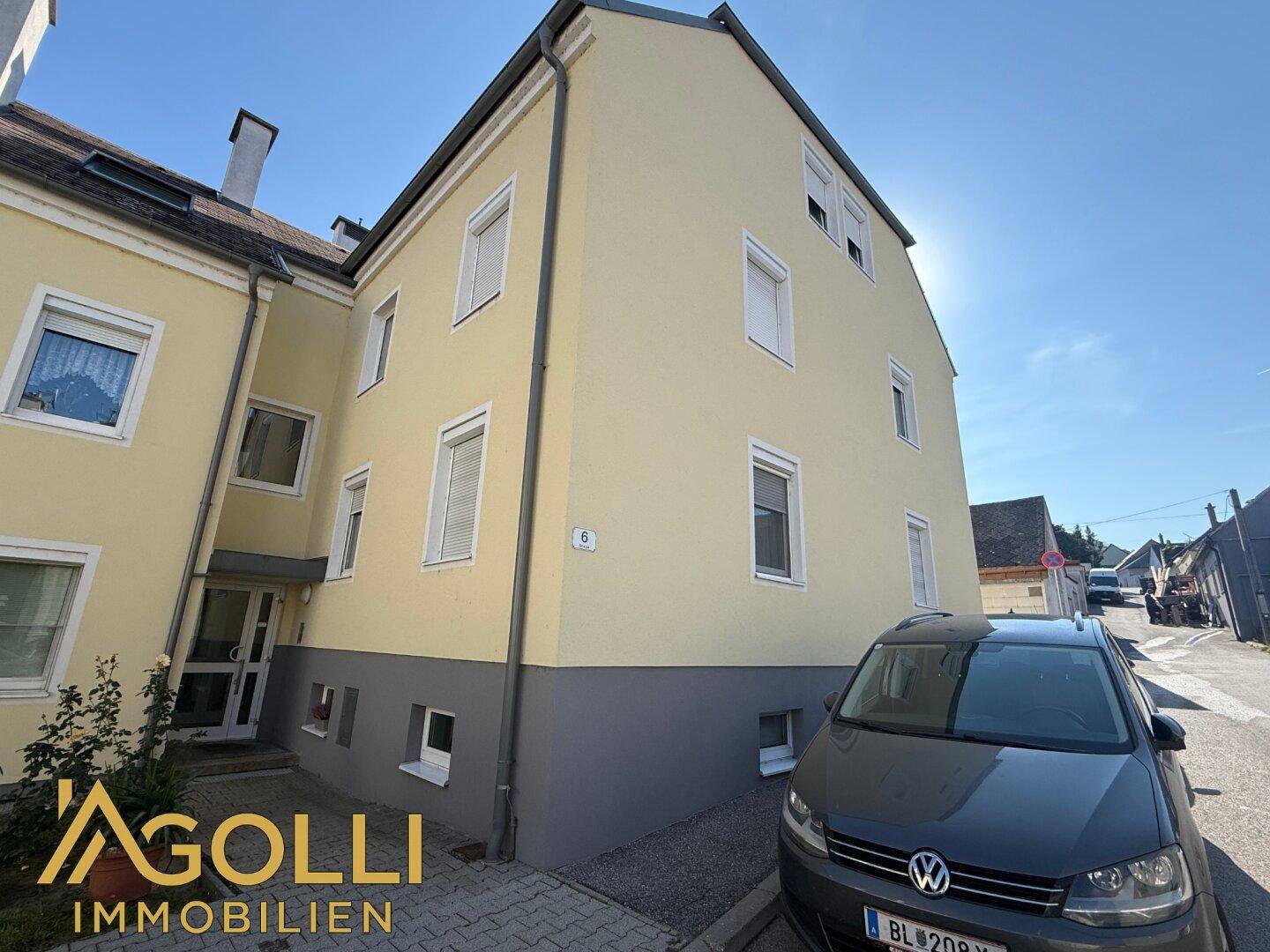 Gelbe Fassade eines Mehrfamilienhauses mit grauen Akzenten, modernen Fenstern und Parkplatz davor.