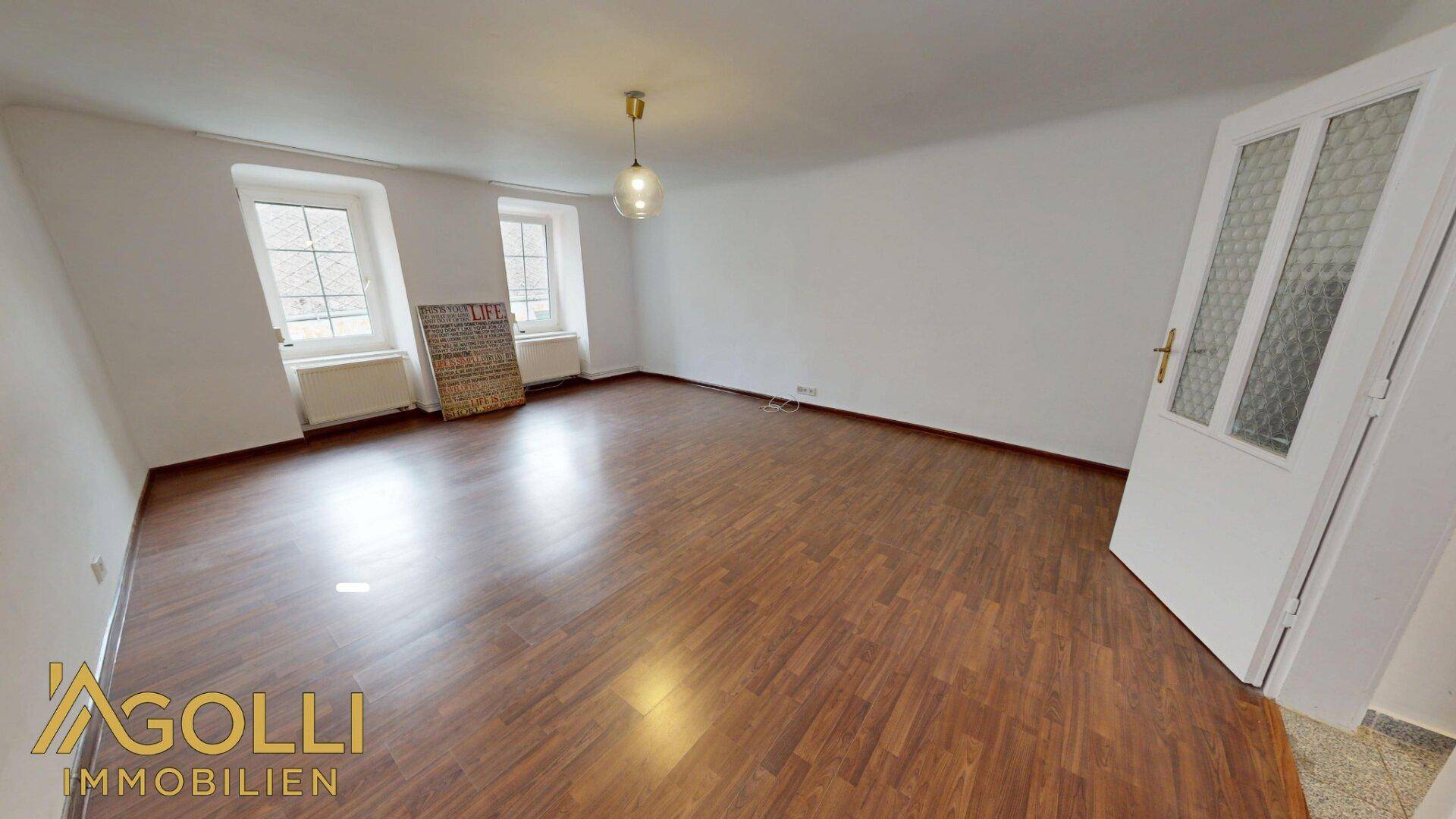 Geräumiges Wohnzimmer mit dunklem Laminatboden und zwei Fenstern, unmöbliert.