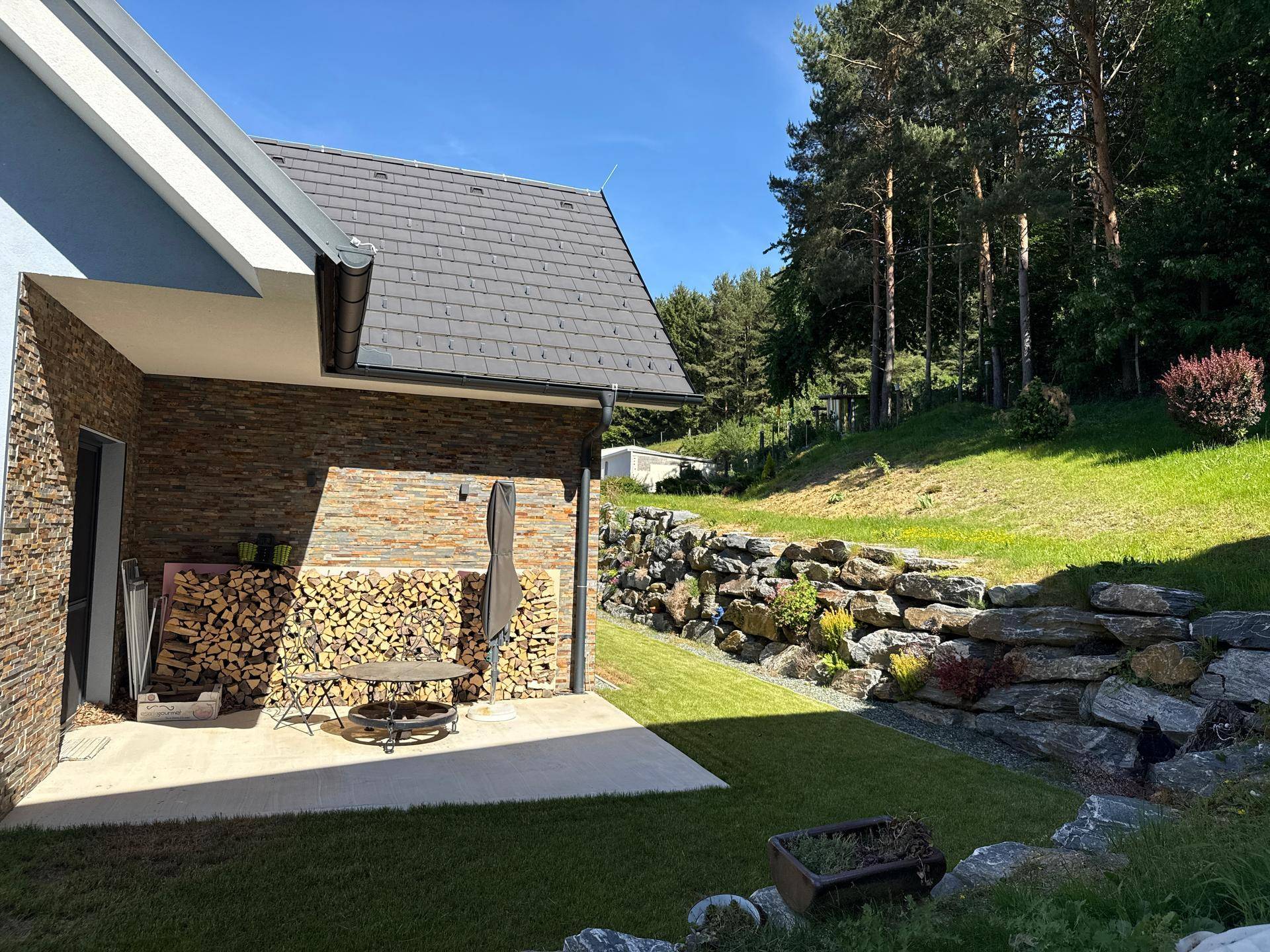 Weitläufiger Garten mit Rasen, Felsformationen und angrenzendem Waldgebiet unter blauem Himmel.