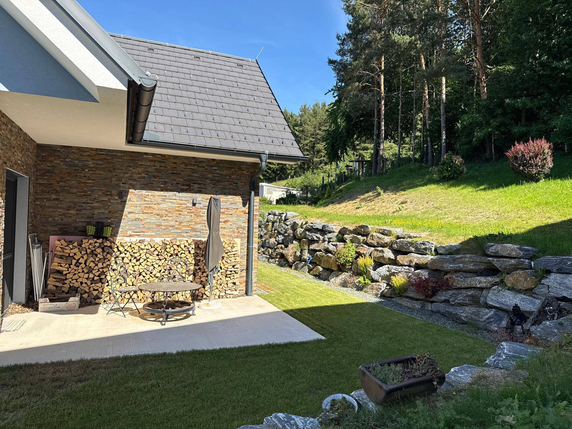 Gepflegter Gartenbereich mit Rasenfläche, dekorativer Felswand und Blick auf bewaldete Hügel.