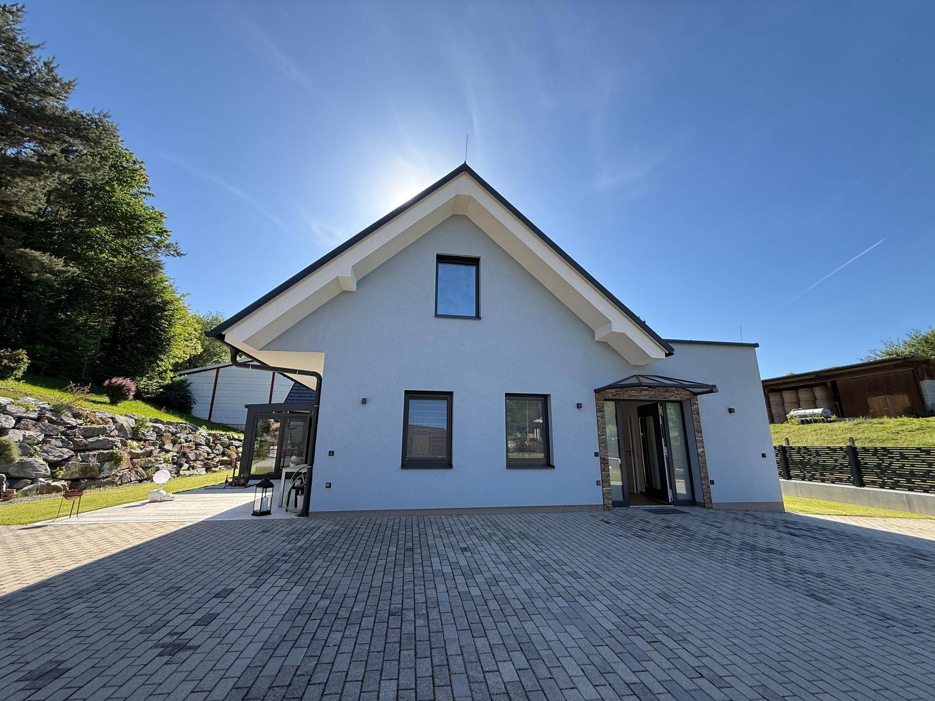 Moderne Hausfassade mit gepflasterter Auffahrt und großem Giebeldach unter strahlendem Sonnenschein.