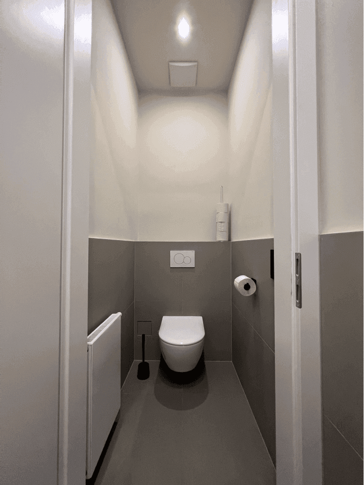 Helles Gäste-WC mit wandhängender Toilette, Heizkörper und grauen Fliesen, minimalistisch und praktisch eingerichtet.
