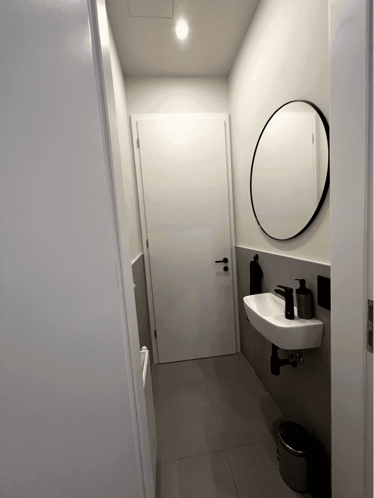 Modernes Gäste-WC mit kleinem Waschbecken, rundem Spiegel und grauen Fliesen, sauber und funktional gestaltet.