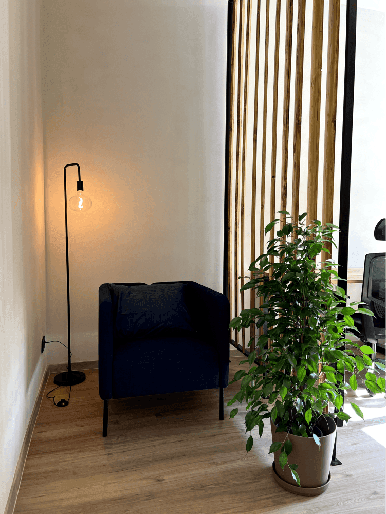 Gemütliche Sitzecke im Büro mit einem blauen Sessel, einer Stehlampe und einer grünen Pflanze neben einer Holztrennwand.