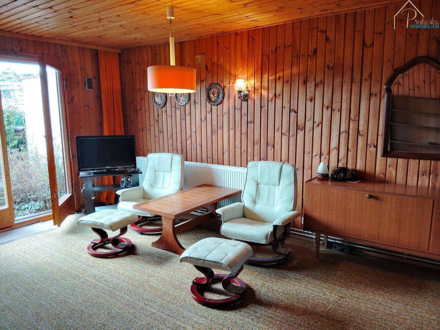 Gemütliches Wohnzimmer mit Holzvertäfelung, bequemen Sesseln und Blick ins Grüne.